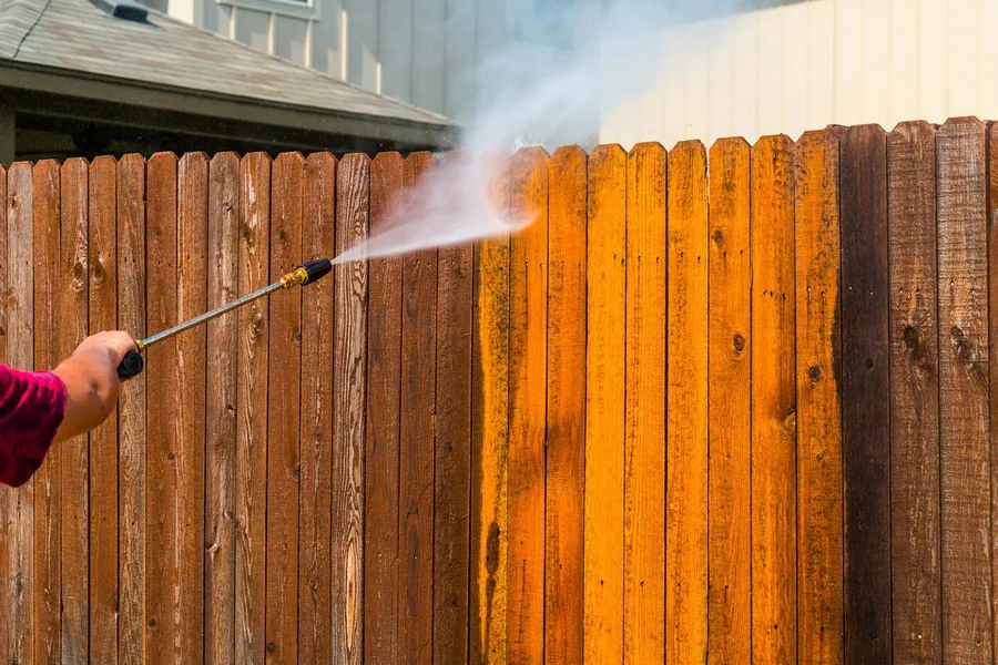 A pressure washer wand stripping grime from board fencing, the cleaned timber bright against the weathered boards
