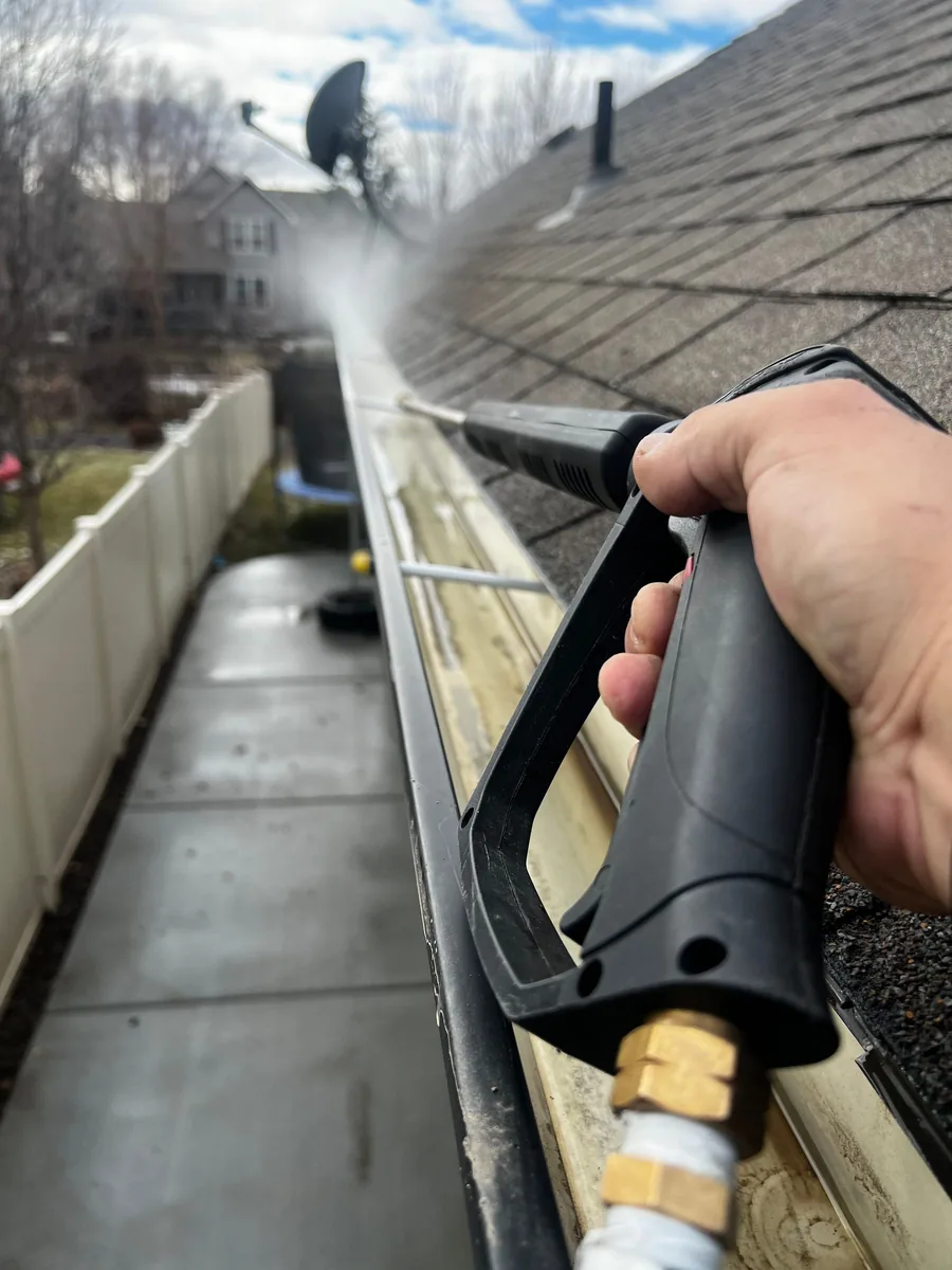 A pressure washer wand rinsing debris out of a gutter along a shingle roof